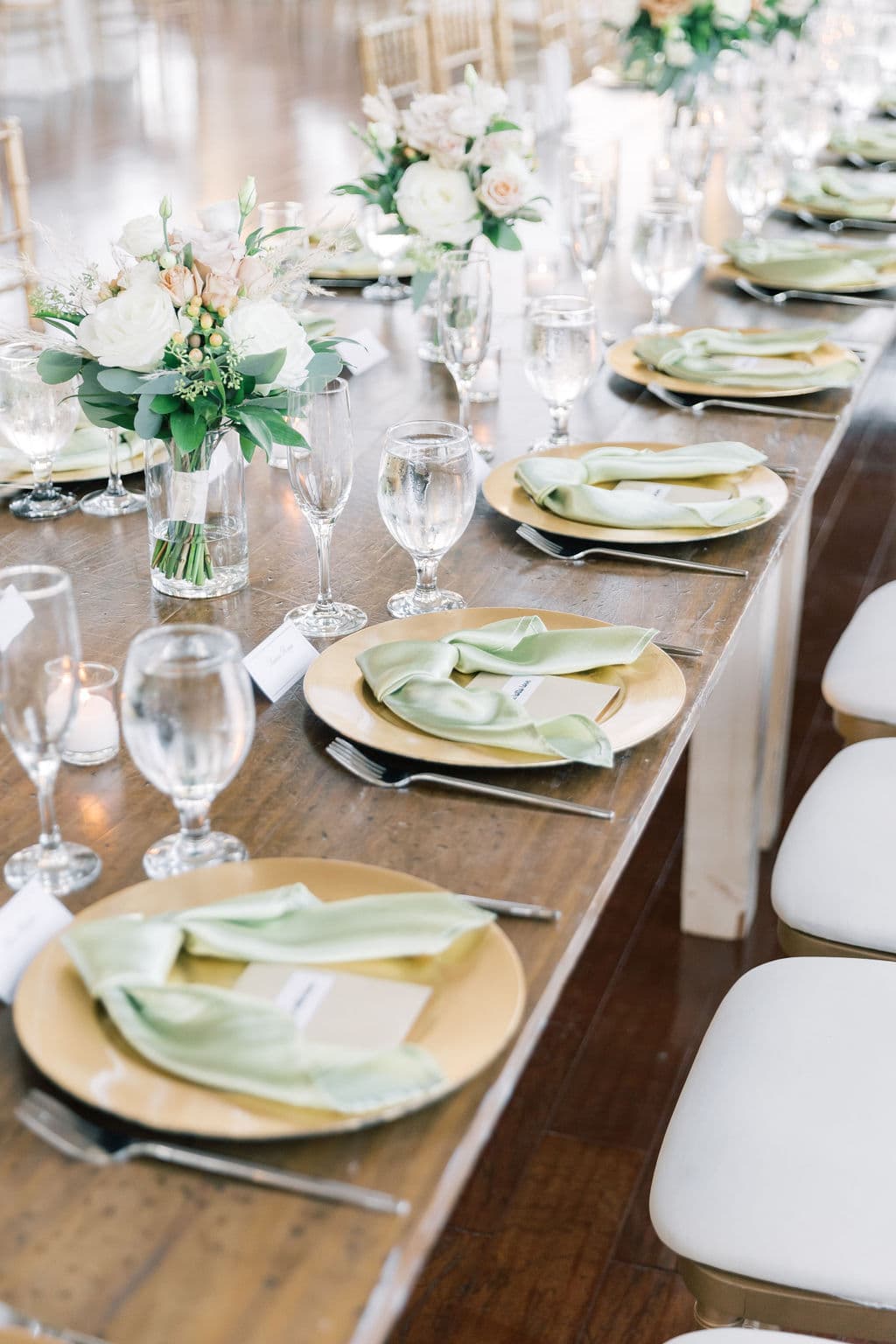 Long farm table set with gold chargers, sage napkins, and white garden roses for a private celebration