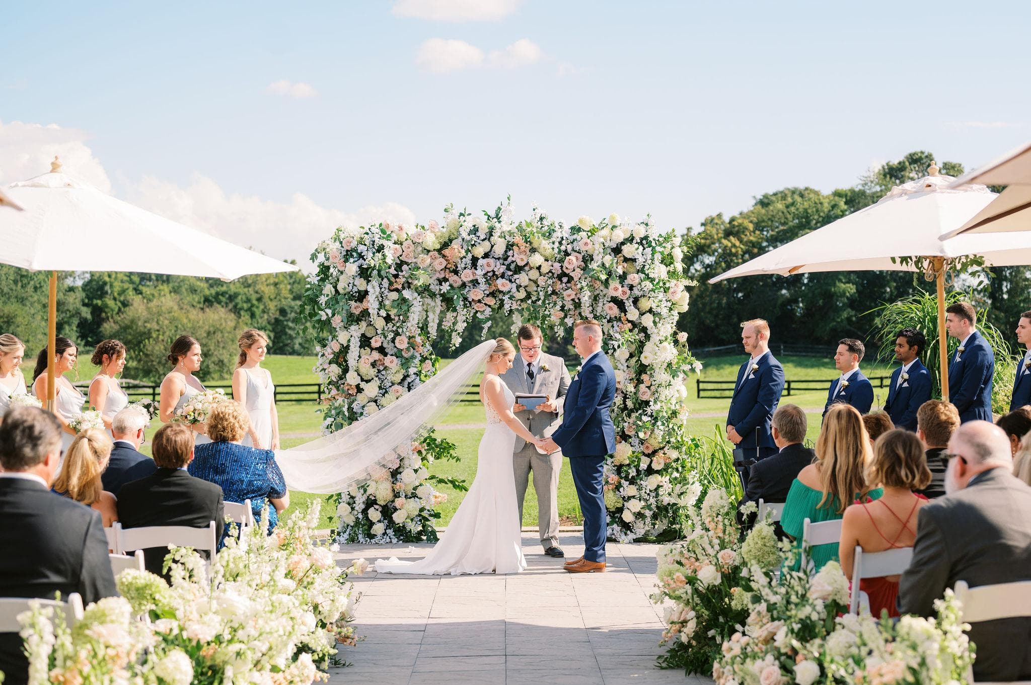 Couple exchanging vows beneath a full floral arch flanked by white umbrellas and their wedding party